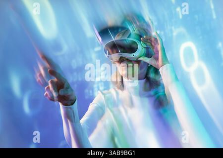 An elderly Asian woman wearing a virtual reality headset immerses herself in the metaverse. She interacts with a futuristic digital environment, showc Stock Photo