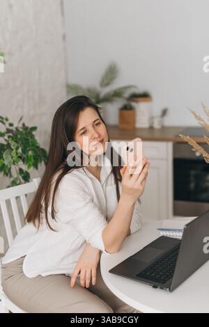 A nutritionist sits at a white desk, engaging in an online consultation via smartphone. A laptop and notebook are on the table, creating a modern home Stock Photo