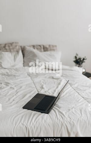 A sleek black laptop rests open on a white bed in a minimalist bedroom. Soft natural lighting and neutral tones create a cozy and modern work-from-hom Stock Photo