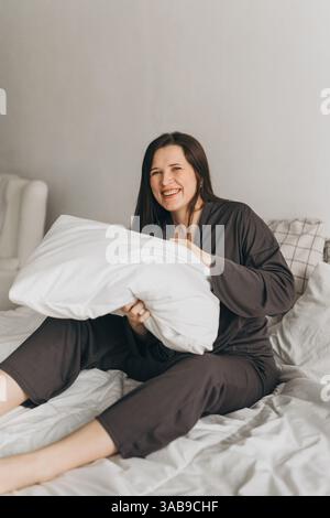 A cheerful young woman in comfortable loungewear sits on a white bed, holding a fluffy pillow with a playful smile. The cozy bedroom setting creates a Stock Photo