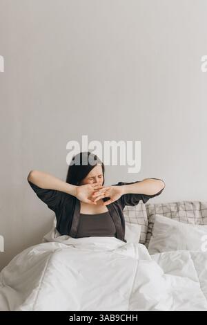 A young woman wakes up and yawns while stretching in bed, wrapped in white blankets. She appears sleepy and relaxed, enjoying a slow morning in a cozy Stock Photo