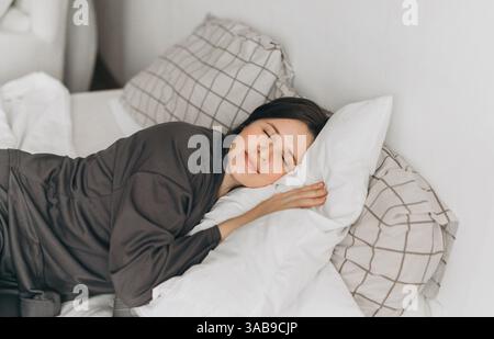 A young woman in comfortable loungewear lies on a white bed, resting her head on soft pillows with a peaceful smile. The cozy bedroom setting creates Stock Photo