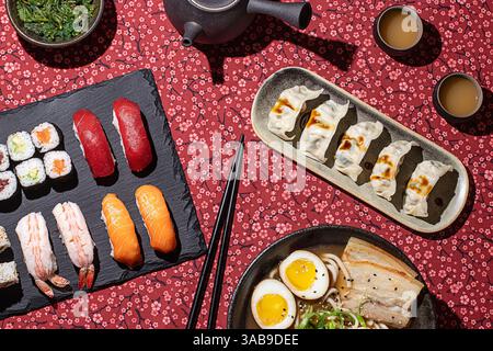 Flat lay of a traditional Japanese meal featuring nigiri, maki rolls, gyoza, and a bowl of ramen, presented on a floral red tablecloth with dramatic s Stock Photo