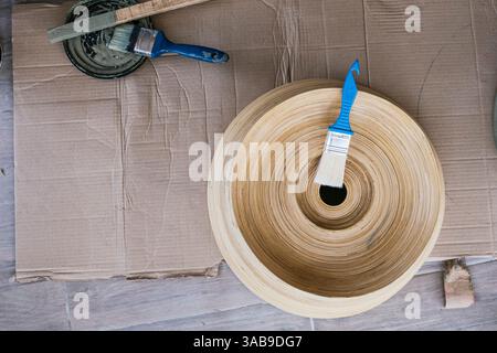 Overhead view of artisan painting tools, including brushes and paint, next to a wooden sculpture placed on a cardboard surface, capturing a rustic and Stock Photo