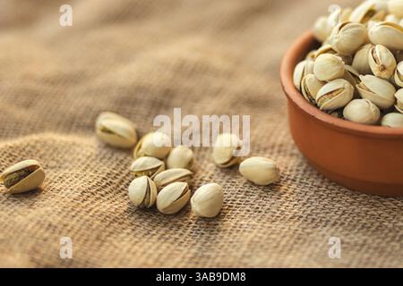 Close up of a clay bowl filled with pistachios on a burlap surface. Several pistachios are scattered around, highlighting the rich texture and warm to Stock Photo