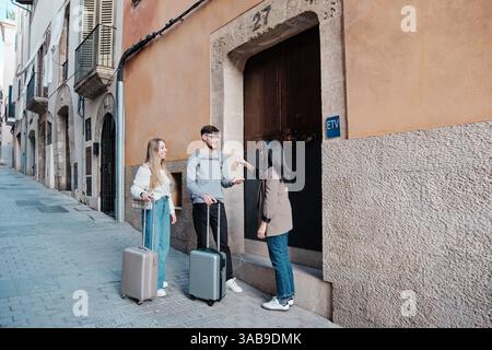 A young couple stands with luggage, receiving keys to their vacation home from a real estate agent. They are on a charming street in Majorca, Spain, e Stock Photo