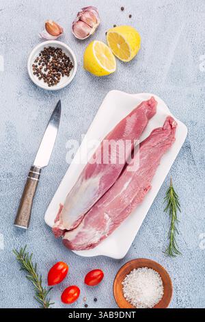 A fresh pork tenderloin on a white tray, surrounded by garlic, lemon, rosemary, tomatoes, salt, pepper, and a knife on a textured surface, ready for c Stock Photo