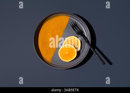 A minimalistic still life features two orange slices and turmeric powder on a round black plate, accompanied by a black fork, creating a striking mono Stock Photo