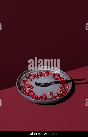 Artistic top view of a dark grey plate with pomegranate seeds arranged in a circular pattern, accompanied by a black fork, set against a rich burgundy Stock Photo