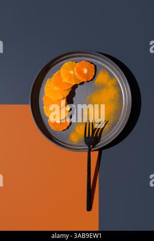 Sliced oranges and tangerines arranged creatively on a gray plate with contrasting orange and dark backgrounds. A black fork complements the minimalis Stock Photo