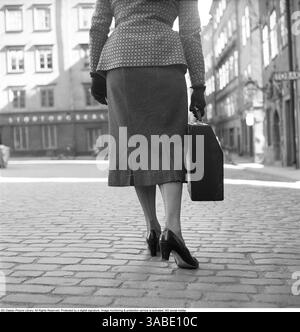 A 1950s woman walking along a cobblestone street in Gamla stan, Sweden, in 1955.  She is dressed elegantly in a patterned jacket, a knee-length skirt, gloves, and high-heeled shoes, reflecting the fashion of the era.    In one hand, she carries a travel typewriter in its designated case, designed for easy transport. This model of typewriter was smaller than a standard one, making it more practical for writers, journalists, and businesspeople who needed to work on the go before the advent of laptops. The photograph captures a glimpse into a past era, where portable typewriters were essential to Stock Photo