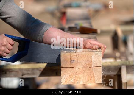 A person uses a hand saw to cut a wooden block on a workbench outside in daylight. Stock Photo