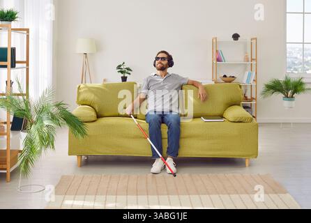 Visually impaired man with cane sitting on couch and listening to music through headphones Stock Photo