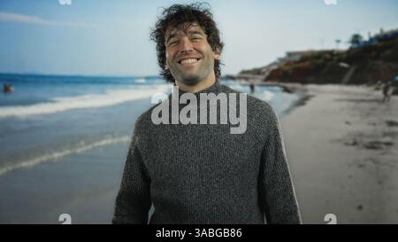 Young man smiling at seaside with wavy hair wearing sweater, exuding happiness and warmth in the serene beach setting under a clear sky, embodying rel Stock Photo