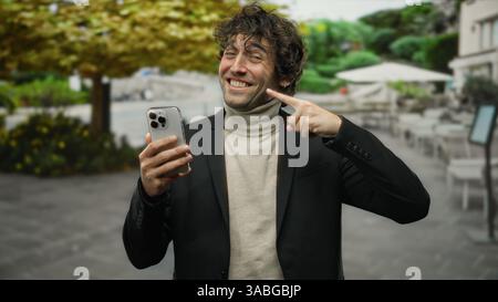 Young man smiling outdoors pointing to his phone while standing in a city street, showcasing confident style and handsome appearance amidst urban scen Stock Photo