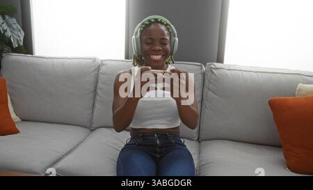 Young woman with braids enjoying game on smartphone in modern living room while wearing headphones and smiling. Stock Photo