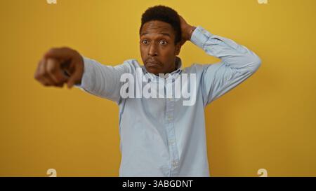 Young man pointing forward with one hand on head against yellow background wearing blue shirt in a comically surprised expression that's striking and Stock Photo