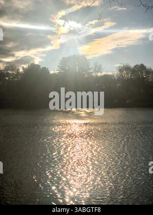A stunning view of the sun peeking through soft, billowy clouds over a tranquil lake. Stock Photo