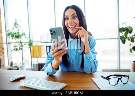 Smiling young businesswoman checking notifications on smartphone while seated in a modern office workspace Stock Photo