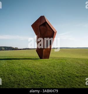 Rusty object in an empty field. Abstract corten steel geometric shape on a lawn mimicking human heart or ear, blue sky in the background. Stock Photo