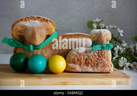 Two Easter lamb cakes with a green ribbon around their necks, three dyed Easter eggs and a sprig of cherry blossoms, gray background, copy space. Stock Photo