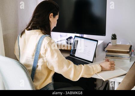 Young Asian woman sitting curled up at her desk, looking at a laptop displaying a ChatGPT interface. Creativity with ChatGPT, digital collaboration Stock Photo