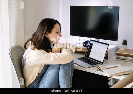 Young Asian woman sitting curled up at her desk, looking at a laptop displaying a ChatGPT interface. Creativity with ChatGPT, digital collaboration Stock Photo