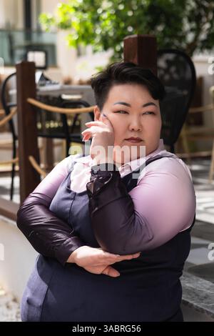 A young plus size Asian woman sits confidently outside a cafe, wearing a stylish outfit. She rests her chin on her hand while gazing off to the side, Stock Photo