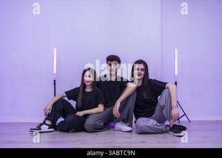 Three young dancers are sitting together on the floor of their dance studio, taking a break and enjoying the purple ambient lighting Stock Photo