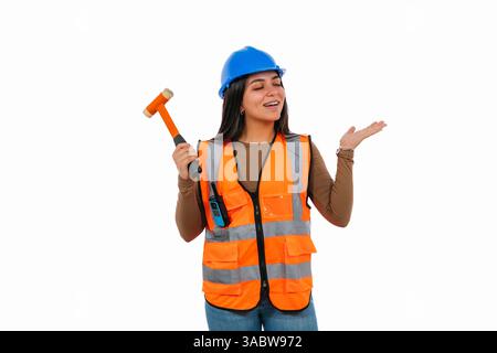 Young smiling Latin woman working as a construction worker, wearing a safety helmet and vest, holding a hammer while gesturing confidently with her ha Stock Photo
