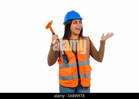 Excited Latina construction worker in an orange safety vest and blue helmet, holding a hammer and gesturing as if presenting against a white backgroun Stock Photo