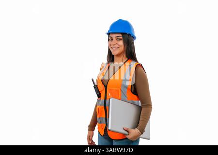 Young latin woman architect or engineer wearing protective workwear and holding laptop and walkie talkie smiling happy while walking on white backgrou Stock Photo