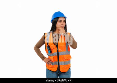 Studio portrait of a thoughtful latin construction engineer with reflective vest, walkie talkie and blue helmet is holding her chin while looking away Stock Photo