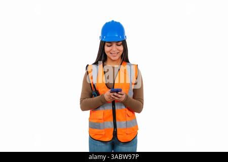 Young construction worker wearing safety vest and helmet using mobile phone, isolated on white background, representing women in construction and tech Stock Photo
