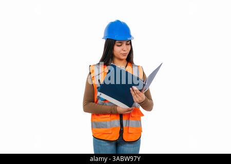 Young Latin construction engineer reading a blueprint while wearing an orange safety vest and a blue helmet, isolated against a clean white background Stock Photo