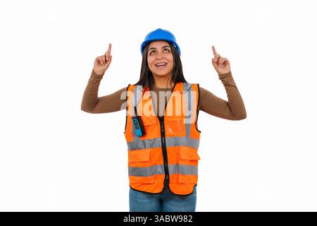 Young construction worker woman wearing safety helmet and vest, pointing up with both index fingers and looking up, smiling, isolated on white backgro Stock Photo