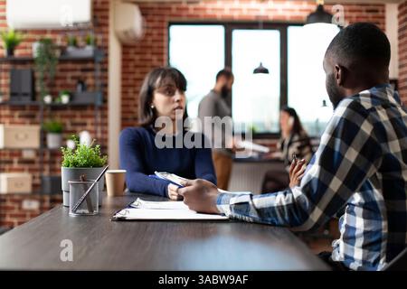 Professional male recruiter is seen assessing female candidate during an employment meeting in brick wall workspace. African american hr manager holding a clipboard and interviewing a white woman. Stock Photo