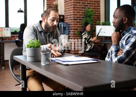 Caucasian man seated at desk, describing his work experiences to african american recruiter. Black hr manager listening to bearded male candidate during job interview in brick wall office. Stock Photo