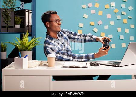 Marketing specialist having a gaming session at his desk, using a gamepad and laptop during his break. Cheerful startup project manager having fun at his workspace, playing video games. Stock Photo