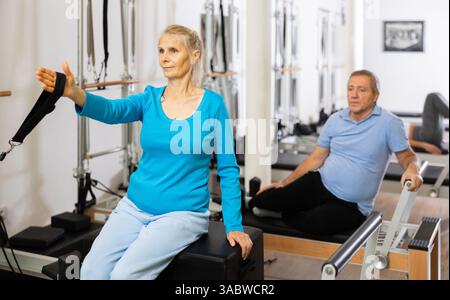 Mature couple using bed of Pilates reformer bed equipment while training together in wellness center. Wellbeing and healthy lifestyle concept Stock Photo
