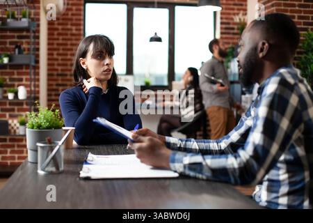 Black male recruiter discussing job options with caucasian female candidate at desk. African american manager advising young woman on company policies during employment interview in modern office. Stock Photo