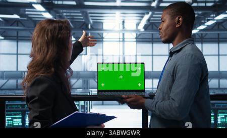 Engineering team in data center using green screen laptop, implementing predictive analytics scripts, reviewing equipment health, adjusting system parameters, coordinating maintenance tasks, camera B Stock Photo