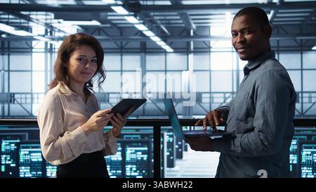 Smiling admins using laptop in data center, managing server virtualization. Cheerful IT specialist and african american man using notebook in server room, ensuring infrastructure reliability, camera B Stock Photo