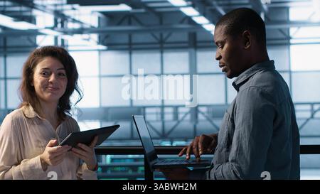 Data scientists in server room using laptop and tablet, executing security patches, verifying firewall configurations. People performing equipment vulnerability scans in data center, camera A Stock Photo