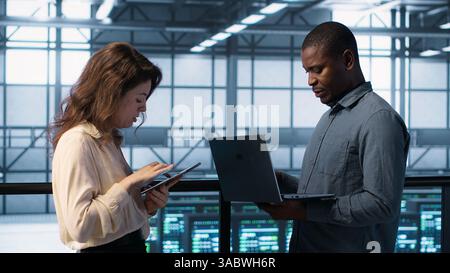 Developers in data center using laptop and tablet, executing security patches, securing firewall. Server farm workers using devices to perform vulnerability scans, ensuring data protection, camera A Stock Photo