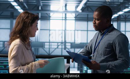 Data center engineer greeting colleague, examining system diagnostics paperwork together. Server room workers doing handshake, meeting to look over maintenance reports, camera A Stock Photo