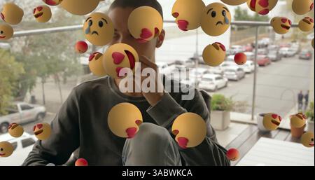 Young man sitting on balcony surrounded by floating emoticons, contemplating life Stock Photo