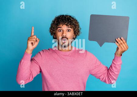 Surprised Young Man Holding Speech Bubble While Pointing Upwards Against Blue Background Stock Photo