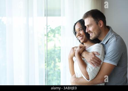 Happy interracial couple hugging looking away through a window standing at home Stock Photo
