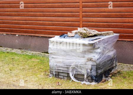 In front of a charming wooden fence, there is a noticeable pile of bricks that have been carefully wrapped in protective plastic sheeting Stock Photo